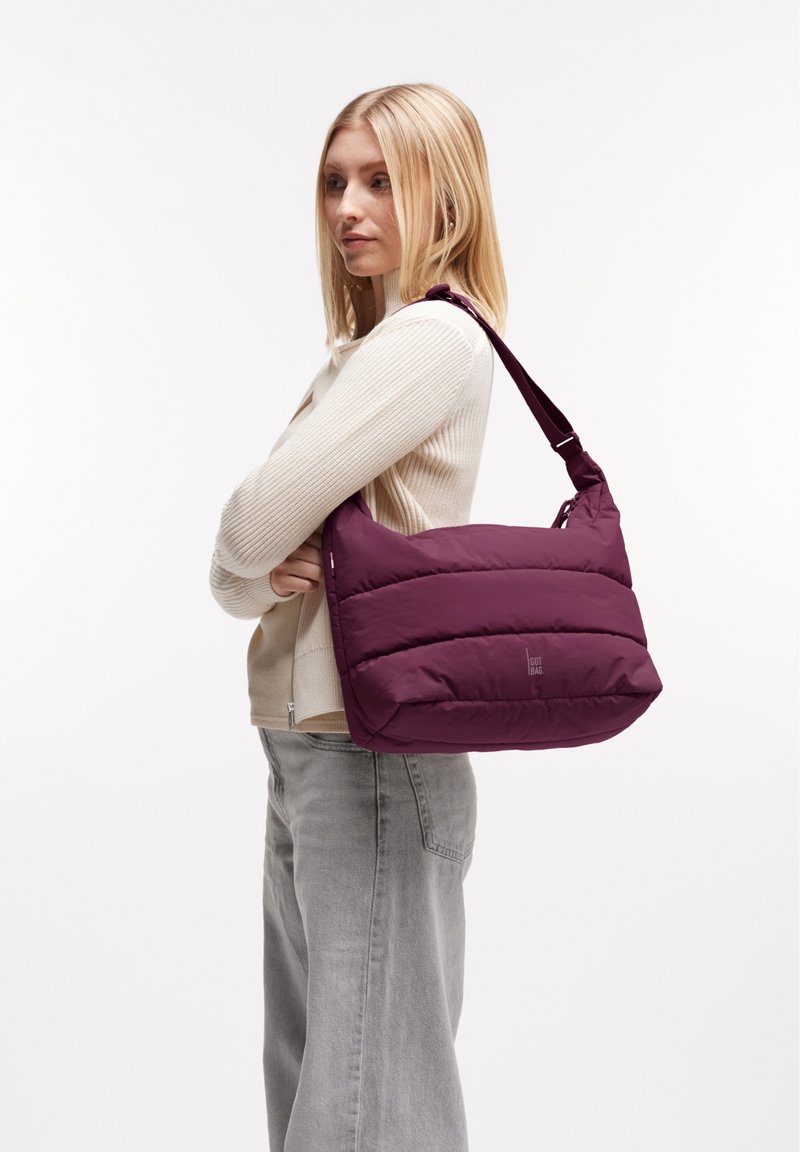 Bolso de hombro morado con un diseño acolchado, textura suave y correa ajustable. Presenta paneles horizontales cosidos y un logo en el lateral.