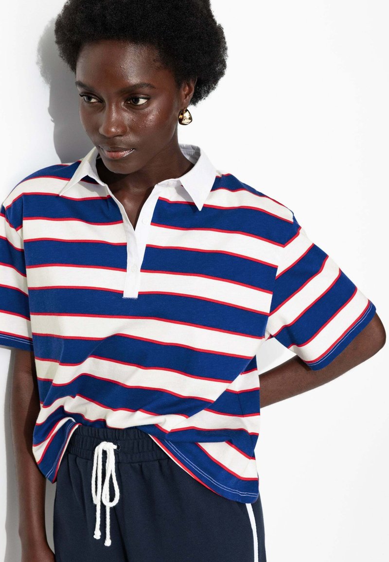 Polo shirt rayé avec des couleurs blanche, bleue et rouge, présentant un col et des manches courtes. Fabriqué en tissu coton doux.