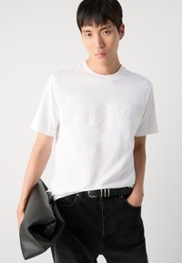 Weißes Kurzarm-T-Shirt mit einem geprägten "BOSS"-Logo. Kombiniert mit schwarzen Jeans und hält ein schwarzledernes Accessoire.