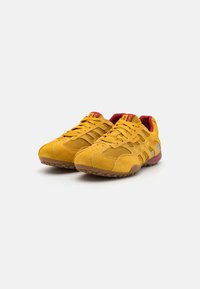Gule semskede sneakers med mesh-detaljer, gummisål, røde accenter og snørebånd. Har en teksturert overdel og sidestriper.