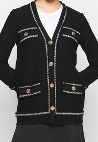 Černý cardigan s texturovaným materiálem, zdobený stříbrným a duhovým prošíváním podél okrajů, dekorativními kapsami a zlatými knoflíky.