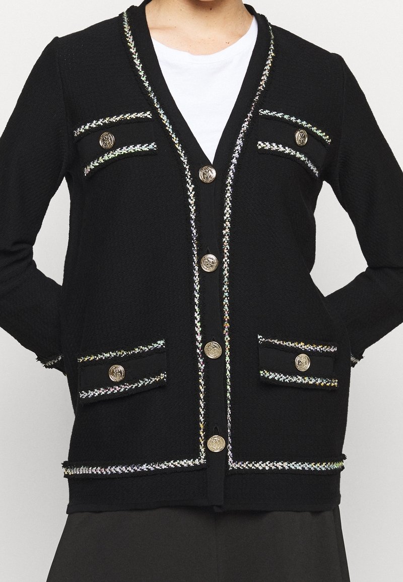Černý cardigan s texturovaným materiálem, zdobený stříbrným a duhovým prošíváním podél okrajů, dekorativními kapsami a zlatými knoflíky.