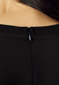 Dettaglio sul retro di un indumento nero con una piccola cerniera e un colletto a coste. Tessuto liscio e morbido con una pelle leggermente visibile sullo sfondo.