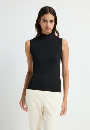 Jeune femme avec de longs cheveux noirs portant un haut sans manches noir à côtes et un pantalon blanc taille haute se tenant devant un fond uni.