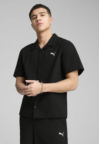 Czarna koszula z krótkim rękawem o teksturowanej tkaninie, z kołnierzykiem w kształcie szpitz i guzikami z przodu. Wyraźne białe logo Puma na piersi.