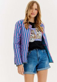 Puchowa kurtka z pionowymi różowymi i niebieskimi paskami, z zamkiem błyskawicznym i ściągaczami przy mankietach. Noszona na czarnej koszulce z grafiką i dżinsowych szortach.