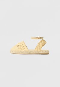 Sandale tressée jaune avec un bout fermé, une sangle �à la cheville réglable et une semelle en jute texturée, avec des motifs découpés sur les côtés.