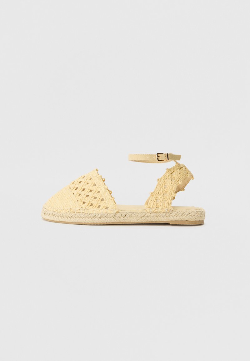 Sandale tressée jaune avec un bout fermé, une sangle à la cheville réglable et une semelle en jute texturée, avec des motifs découpés sur les côtés.