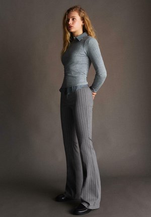 Mlada ženska z dolgimi blond lasmi, oblečena v siv oprijet pulover, črtasto srajco, sivim hlačami s tankimi črtami in črnimi čevlji, stoji bočno.