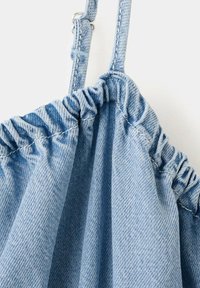 Tissu en denim bleu clair avec bord élastique froncé et sangle réglable avec boucle métallique sur fond blanc.