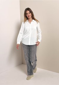 Chemise blanche à manches longues avec boutons portée avec un pantalon gris. La tenue présente un col classique et une coupe décontractée, sur fond neutre.