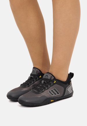 Piedi con indosso scarpe da ginnastica leggere nere e grigie con suole testurizzate e accenti gialli su sfondo bianco.