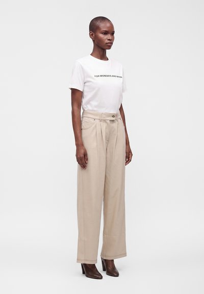 Femme portant un t-shirt blanc avec un texte, un pantalon beige à jambes larges et des chaussures marron à talons hauts, debout devant un fond uni.