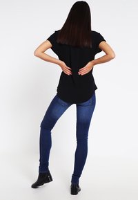 Sort kortærmet top med buet kant, sat sammen med tætsiddende blå jeans og sorte ankelstøvler, set bagfra.
