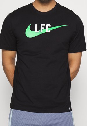 Mann trägt ein schwarzes T-Shirt mit grünem Nike-Swoosh und weißen Buchstaben "LFC" auf der Brust, kombiniert mit blauen gemusterten Shorts.