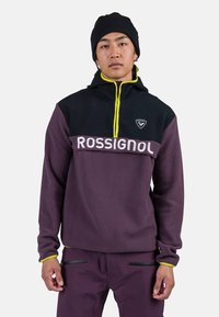 Pullover in pile di colore viola scuro e nero, con zip gialla e logo "ROSSIGNOL" sul petto. Tessuto morbido con maniche lunghe.