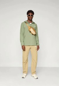 Ανοιχτό πράσινο τζάκετ με φερμουάρ, μπεζ cargo παντελόνι, μπεζ τσάντα χιαστί, λευκά αθλητικά παπούτσια και λευκά γυαλιά ηλίου σε ανδρικό μοντέλο.