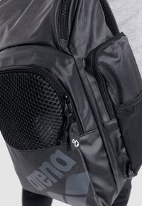 Zaino nero realizzato in tessuto resistente, dotato di tasca in rete, molteplici scomparti con zip e logo prominente sulla parte anteriore inferiore.