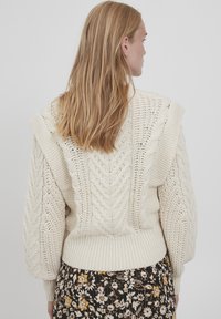 Maglione chunky in maglia con motivi a treccia e maniche puff. Orlo e polsini a coste, indossato sopra una gonna con stampa floreale.