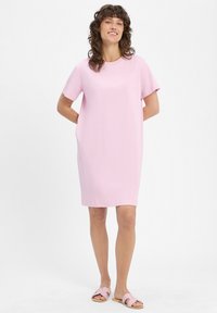 Rosa kurzes Kleid mit kurzen Ärmeln aus weichem Stoff. Rundhalsausschnitt, lockere Passform, knielang. Kombiniert mit hellrosa Slipper-Sandalen, schlichtes Design.