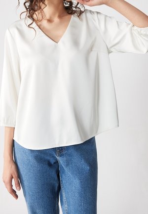 Vrouw draagt een losse witte V-hals blouse met driekwart mouwen, gecombineerd met blauwe denim jeans, arm omhoog dichtbij het gezicht.