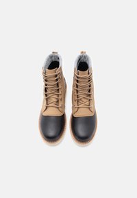 O pereche de cizme de iarnă bej și negre, cu șireturi și căptușeală din polar gri, văzută dintr-o parte de sus pe un fundal simplu.