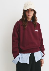 Bordeaux sweatshirt met geribbelde zoom, voorzien van witte tekst "LOS ANGELES". Gecombineerd met een blauw gestreept shirt en een beige pet met tekst.