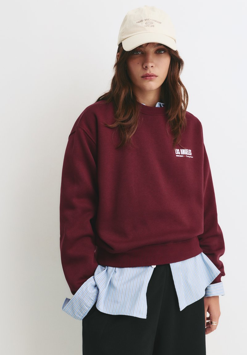 Bordeaux sweatshirt met geribbelde zoom, voorzien van witte tekst "LOS ANGELES". Gecombineerd met een blauw gestreept shirt en een beige pet met tekst.