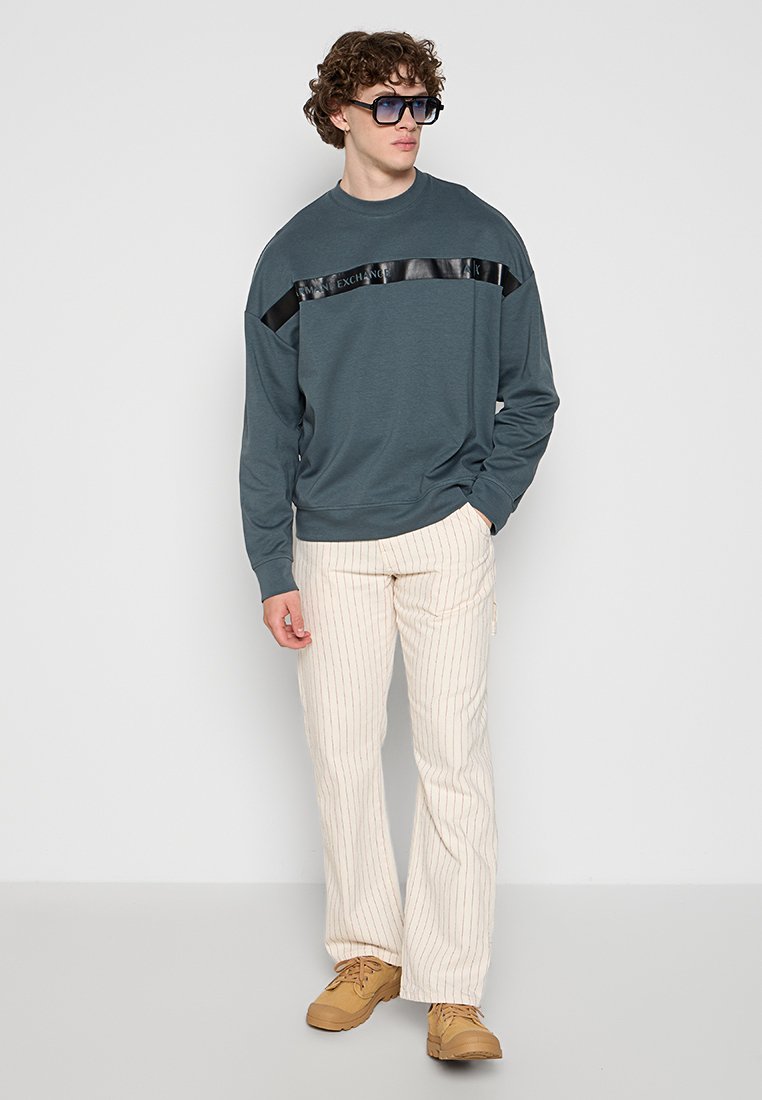 Junger Mann in einem dunkelgrauen Sweatshirt mit schwarzem Streifen, cremfarbenen Hosen mit dünnen roten Streifen, hellbraunen Schuhen und großen dunklen Sonnenbrille, der an einer Wand steht.