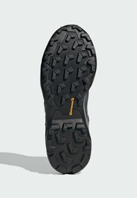 Semelle de chaussure noire avec un motif de semelle robuste, comprenant des rainures inclinées et des sections surélevées pour une meilleure traction. Comprend un petit logo orange.