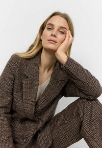 Brun tweed-blazer med en struktureret vævning, der har en skrå krave og tre knapper på manchetterne, parret med en beige top.