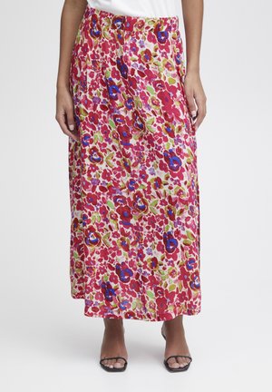 Femme portant une longue jupe fleurie colorée avec des motifs rouges, bleus et verts, associée à des sandales noires ouvertes à l'avant et un haut blanc.