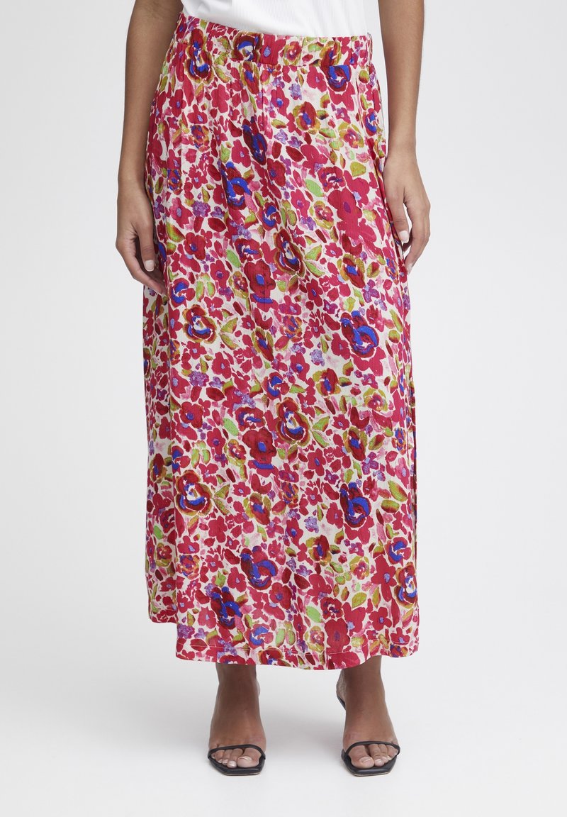 Femme portant une longue jupe fleurie colorée avec des motifs rouges, bleus et verts, associée à des sandales noires ouvertes à l'avant et un haut blanc.