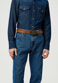 Camisa de mezclilla con botones a presión, dos bolsillos en el pecho y detalles en el yugo al estilo western, combinada con jeans azules y un cinturón de cuero marrón con una hebilla de metal.