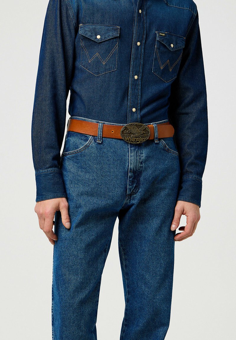 Denimskjorte med trykknapper, to brystlommer og western-yoke detaljer, parret med blå jeans og et brunt læderbælte med en metalspænde.