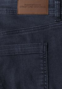 Jeans de mezclilla azul oscuro con costura detallada y una etiqueta de cuero marrón que dice "diseñado con pasión STREET ONE DENIM ATELIER."
