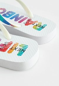 Des tongs blanches avec des semelles texturées, ornées d'inscriptions multicolores "NEXT" et de graphiques à la main en bleu, violet, rouge et vert.
