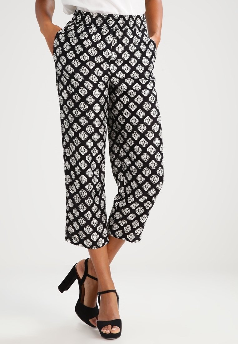 Femme portant un pantalon court noir à motif floral blanc et des sandales noires à talons hauts, mains dans les poches.