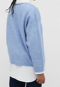 Hellblauer Pullover aus weichem Strickstoff, mit lockerer Passform, geripptem Saum und langen Ärmeln, die über einem weißen Hemd getragen werden.