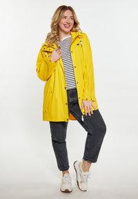 Geltona vandeniui atspari striukė su kapu, užsegama spaudėmis, su šoninėmis kišenėmis, dėvima ant juodai baltos juostuotos marškinėlių ir derinama su tamsiomis džinsomis bei sportbačiais.