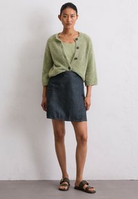 Cardigan vert en peluche avec trois boutons, associé à une jupe en denim foncé avec une poche latérale, et des sandales à glissière marron.