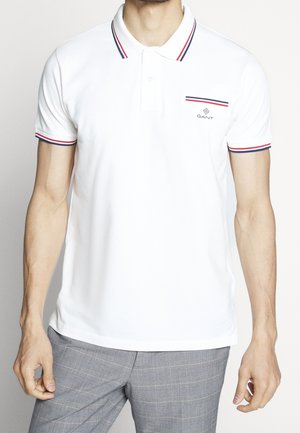 Vit bomullspolo med röd och blå randig krage, med en liten ficka och logotyp på bröstet. Kortsleeves och klassisk passform.