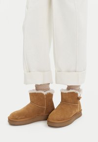 Botine din suede bej cu căptușeală din blană albă, vârf rotund, talpă din cauciuc, cusături decorative și o etichetă mică pe lateral, asortate cu pantaloni albi răsuciți.