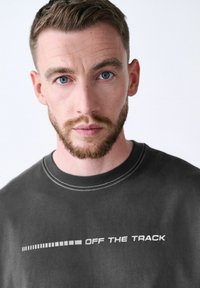 Fekete pamut póló kerek nyakkal, fehér "OFF THE TRACK" felirattal és grafikus mintázattal az alján. Simább textúra.