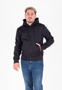 Felpa nera con zip e tessuto testurizzato, tasche anteriori e cappuccio con cordino. Cerniere e dettagli metallici completano il design.