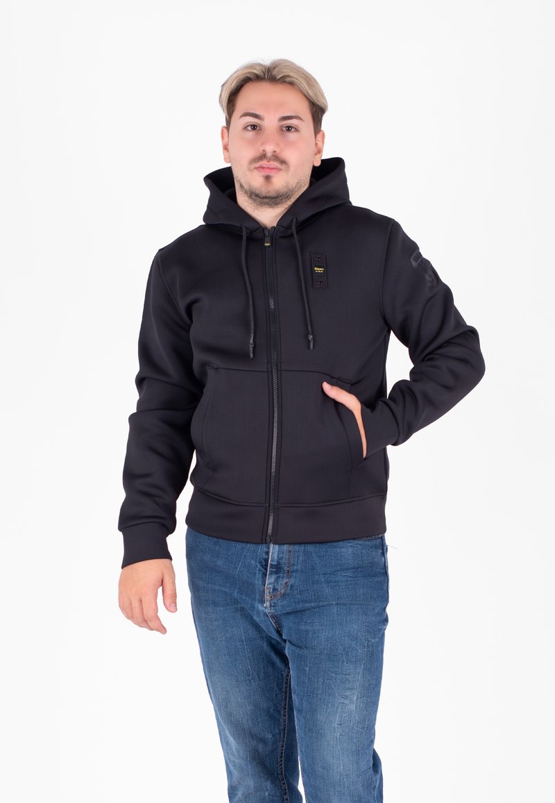 Felpa nera con zip e tessuto testurizzato, tasche anteriori e cappuccio con cordino. Cerniere e dettagli metallici completano il design.