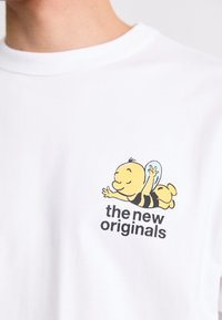 Hvit bomull t-skjorte med en tegneseriebi-karakter i gult og svart, med teksten "de nye originalene" trykket under.