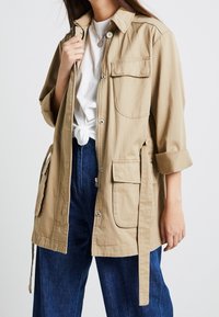 Beige lichtgewicht jack met een kraag, voorzakken, knoopsluiting en verstelbare banden in de taille. Draagt over een wit shirt en blauwe broek.