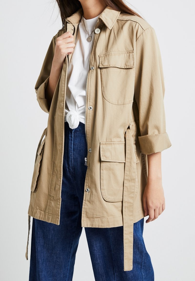 Beige lichtgewicht jack met een kraag, voorzakken, knoopsluiting en verstelbare banden in de taille. Draagt over een wit shirt en blauwe broek.