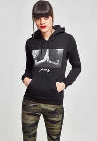 Sudadera negra con capucha y bolsillo frontal, que presenta un gráfico de manos en oración. Combinada con mallas de camuflaje.